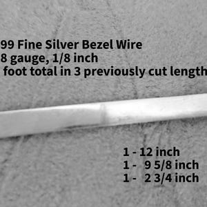 May include: 999 Fine Silver Bezel Wire, 28 gauge, 1/8 inch, 2 foot total in 3 previously cut lengths: 1-12 inch, 1-9 5/8 inch, 1-2 3/4 inch. A single strip of flat, metallic silver wire is shown horizontally against a textured background.