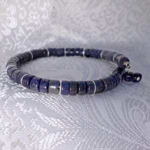 Puede incluir: Una pulsera de cuentas azul oscuro con espaciadores plateados. La pulsera está hecha de pequeñas cuentas cilíndricas y se muestra sobre una superficie clara estampada. Es una pieza de joyería.