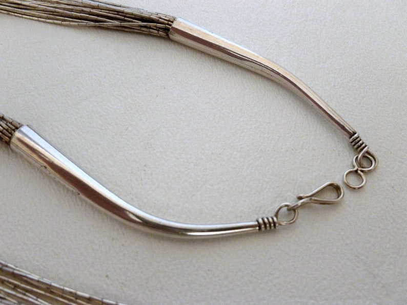 May include: A silver necklace with a chain made of multiple strands of thin silver wire. The necklace has a simple, curved clasp.