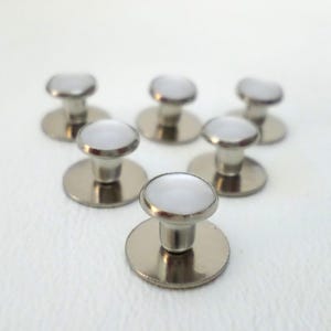 May include: Six silver-toned shirt studs with white, circular tops. The studs are arranged in a semi-circular pattern against a white background. The studs have a classic, minimalist design.