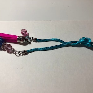 May include: A teal blue cord with a knot, a pink tassel, a blue sparkly bead, and a silver bell charm.