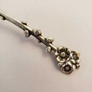 May include: A silver-toned floral spoon with a branch-like handle. The spoon features detailed flower blossoms and buds. The spoon is likely made of sterling silver or a similar metal. The design is elegant and nature-inspired.