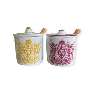 May include: Two white ceramic jars with lids and wooden spoons. One jar has a yellow design, the other has a red design. Both jars have a crest design and the text "QUE MAILLE QUI MAILLE".