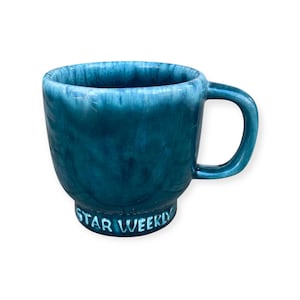 May include: A teal ceramic mug with a rounded body and a handle. The mug has a glossy finish and the words "STAR WEEKLY" are printed on the base. The mug is set against a white background.