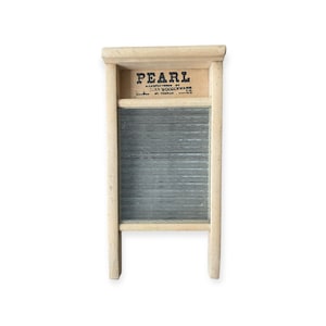 May include: A vintage wooden washboard with a corrugated metal scrubbing surface. The top section features the word "PEARL" in bold letters, with additional text below. The washboard has a light wood frame and a classic design.