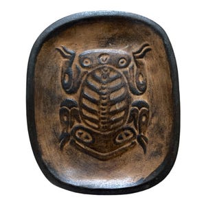 May include: A brown ceramic dish with a black frog design. The frog is stylized and has a detailed design.