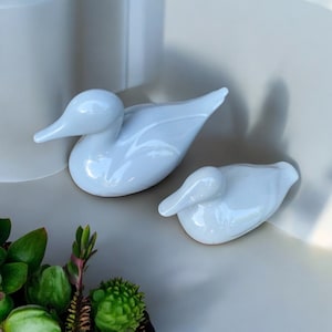 May include: Two glossy white ceramic duck figurines. One duck is upright, the other is lying down. The ducks have smooth, rounded bodies and long, pointed beaks. The figurines are displayed on a neutral-coloured surface.