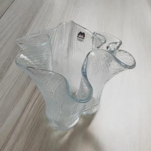 May include: A clear glass bowl with a textured surface and a wavy, ruffled rim. The bowl has a label that reads "movia". The bowl is likely used for decorative purposes.
