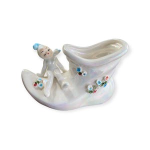 May include: A vintage, pearlescent white ceramic boot-shaped planter with a reclining figurine. The boot is decorated with small, colourful floral accents. The figurine has a blue cap and is in a relaxed pose.