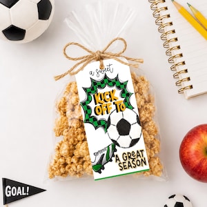 May include: A clear plastic bag filled with caramel popcorn, tied with a brown twine bow. A rectangular white tag features a green and black checkered burst, a soccer ball, a soccer cleat, and the text A SWEET KICK OFF TO A GREAT SEASON.