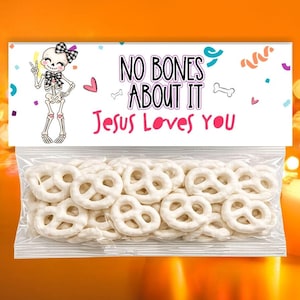 May include: A clear plastic bag filled with white yogurt-covered pretzels, topped with a white header card featuring a cartoon skeleton wearing a black and white plaid bow, confetti, and the text "NO BONES ABOUT IT Jesus Loves YOU".