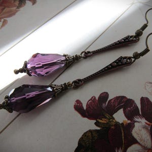 May include: A pair of long drop earrings featuring faceted, teardrop-shaped purple crystals suspended from antique bronze-toned metal components. Each earring includes a decorative metal cap at the top of the crystal and a slender, textured metal bar.