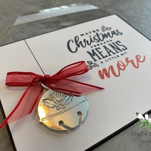 May include: A white card with black text that reads "Maybe Christmas Perhaps Means A Little Bit More". A silver bell ornament with a red ribbon is attached to the card.