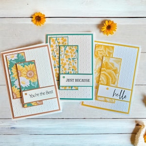 May include: Three handmade greeting cards with floral designs and text. The cards feature a white dotted background with rectangular panels of sunflowers and yellow flowers. Text includes "You're the Best", "Just Because", and "hello".