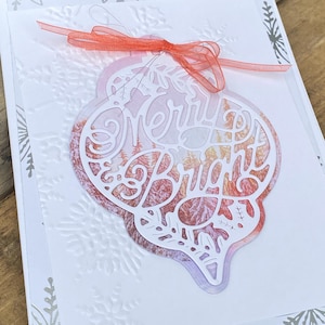 Puede incluir: Una tarjeta de Navidad blanca con un diseño de copo de nieve en relieve y detalles plateados. Un adorno decorativo con las palabras "Merry & Bright" está sujeto con una cinta roja. El adorno tiene un diseño recortado con un fondo colorido.