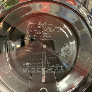 May include: A clear glass pie plate with a handwritten recipe for Mom's Apple Pie. The recipe includes 5-6 apples, 1 tablespoon of lemon juice, 1/2 cup of white sugar, 2 tablespoons of brown sugar, and 2 teaspoons of minute tapioca. The recipe also includes 2 tablespoons of powdered cinnamon. The recipe instructs to mix all ingredients, put in unbaked pie crust, dot with butter, cover with pastry, bake at 425 degrees Fahrenheit for 15 minutes, then reduce heat to 350 degrees Fahrenheit and bake for 30-45 minutes.