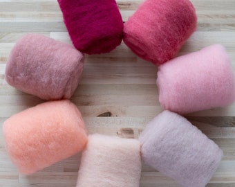 Peut inclure: Sept pelotes de laine de différentes nuances de rose, allant d'un magenta foncé à un blush pâle, disposées en cercle sur une surface en bois clair.
