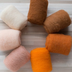 May include: Seven rolls of wool yarn in various shades of orange, brown, and beige. The yarn is arranged in a circle on a light wood surface.