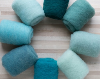 May include: A set of eight rolled up balls of wool yarn in shades of blue and green. The yarn is arranged in a circle on a light wood surface.