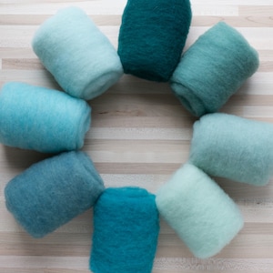 May include: A set of eight rolled up balls of wool yarn in shades of blue and green. The yarn is arranged in a circle on a light wood surface.