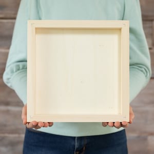 Puede incluir: Una persona sostiene un marco de imagen de madera clara simple. El marco es cuadrado y tiene un diseño simple.