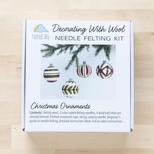 Op de afbeelding: Een witte doos met een naaldviltset om kerstversieringen te maken. De doos heeft een afbeelding van vier vilten ornamenten met verschillende ontwerpen. De tekst op de doos luidt "Decorating With Wool Needle Felting Kit" en "Christmas Ornaments".