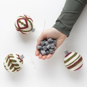 May include: Four felt Christmas ornaments, two with red and green stripes, one with a green and red geometric pattern, and one with a green leaf pattern. A hand holds a pile of gray felt balls.