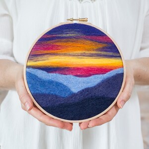 May include: A circular needlepoint embroidery hoop with a sunset scene over a mountain range. The sky is a gradient of purple, orange, and yellow, with a few wispy clouds. The mountains are a deep blue and purple.