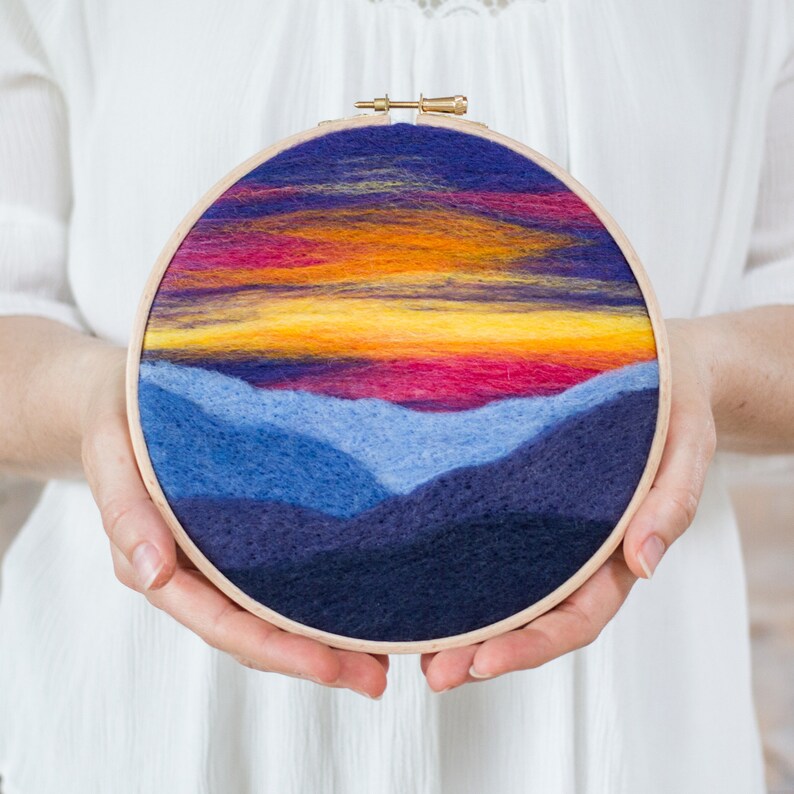 May include: A circular needlepoint embroidery hoop with a sunset scene over a mountain range. The sky is a gradient of purple, orange, and yellow, with a few wispy clouds. The mountains are a deep blue and purple.