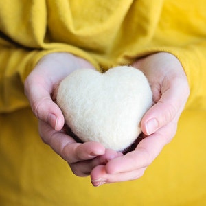 May include: A pair of hands holding a white felt heart. The hands are against a yellow background.