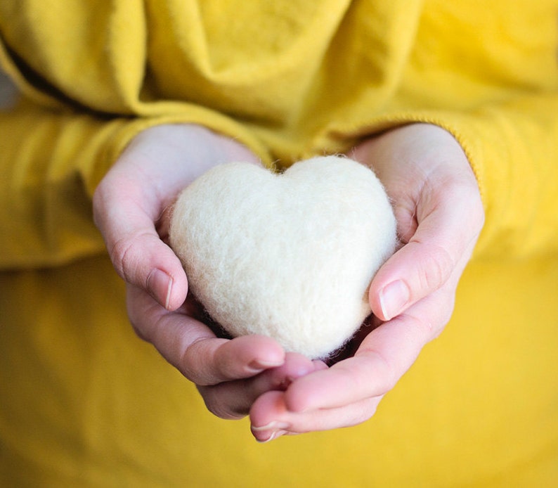 May include: A pair of hands holding a white felt heart. The hands are against a yellow background.