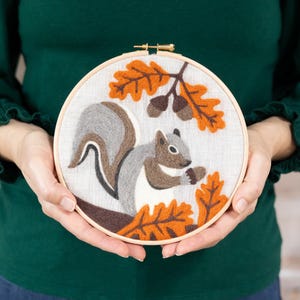 May include: A gray and brown squirrel embroidered on a white fabric with orange and brown leaves and acorns. The embroidery is in a wooden hoop.