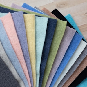 May include: A collection of 18 felt squares in various colors, including light blue, pink, yellow, green, and gray. The felt squares are arranged in a fan shape, with the tips of the squares pointing towards the top of the image.