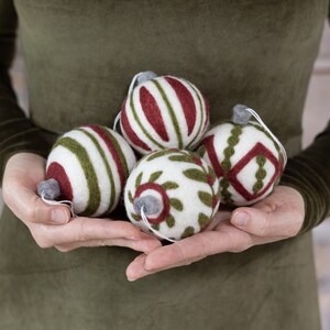 Op de afbeelding: Vijf handgemaakte vilten kerstballen in verschillende patronen. De kerstballen zijn wit met groene en rode details. De kerstballen worden in de handen van een persoon gehouden.