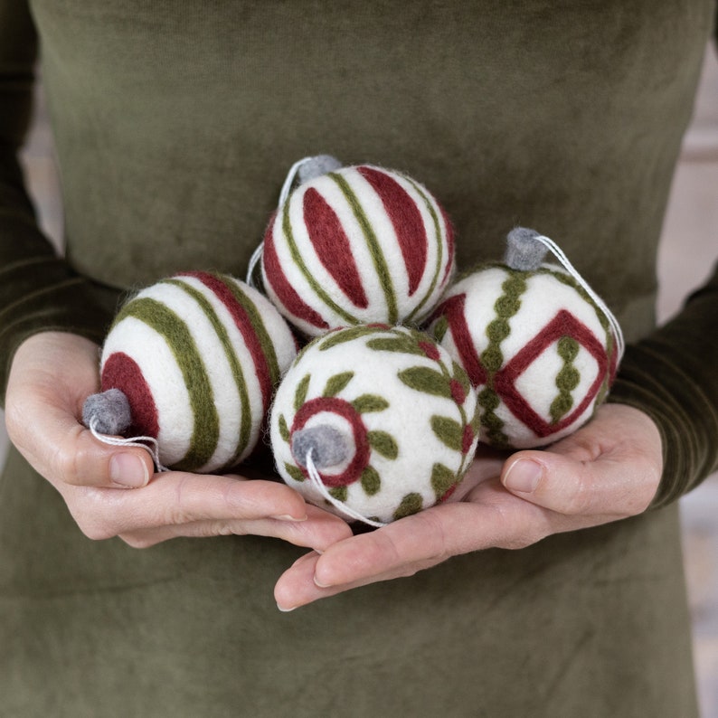 Op de afbeelding: Vijf handgemaakte vilten kerstballen in verschillende patronen. De kerstballen zijn wit met groene en rode details. De kerstballen worden in de handen van een persoon gehouden.