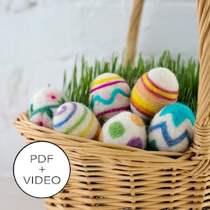 May include: A wicker basket filled with colourful felt Easter eggs. The eggs are decorated with stripes, dots and swirls. The basket is sitting on a white surface.
