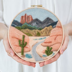 Puede incluir: Un aro circular de bordado de aguja con una escena de paisaje desértico. La escena presenta una formación rocosa roja, un camino sinuoso y dos cactus verdes. El fondo es un cielo azul con montañas grises.