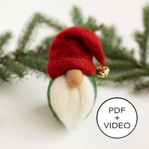 Puede incluir: Un adorno de gnomo de fieltro con un gorro rojo, barba blanca y cuerpo verde. El gnomo sostiene una pequeña campana dorada. El adorno está sobre una superficie blanca con una rama de pino en el fondo. El texto "PDF + VIDEO" es visible en un círculo.