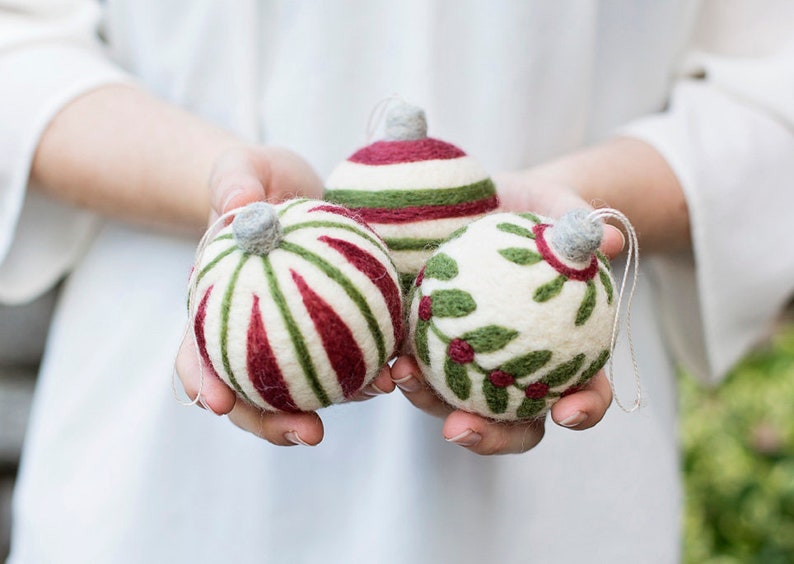 May include: Three handmade felt Christmas ornaments in white, green, and red. The ornaments are decorated with stripes and holly.