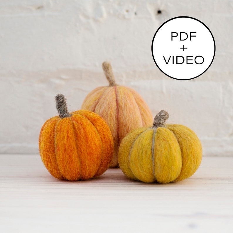 May include: Three handmade felt pumpkins in varying shades of orange and yellow. Each pumpkin has a textured surface and a gray stem. The pumpkins are arranged on a light-colored wooden surface, with a white brick wall in the background.