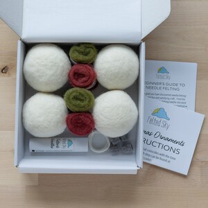 Op de afbeelding: Een witte doos met een wit deksel dat open is, waardoor een naaldviltset voor het maken van kerstversiering zichtbaar wordt. De set bevat witte wollen ballen, groene en rode viltwol, een naaldviltgereedschap en instructies. De doos staat op een houten oppervlak.