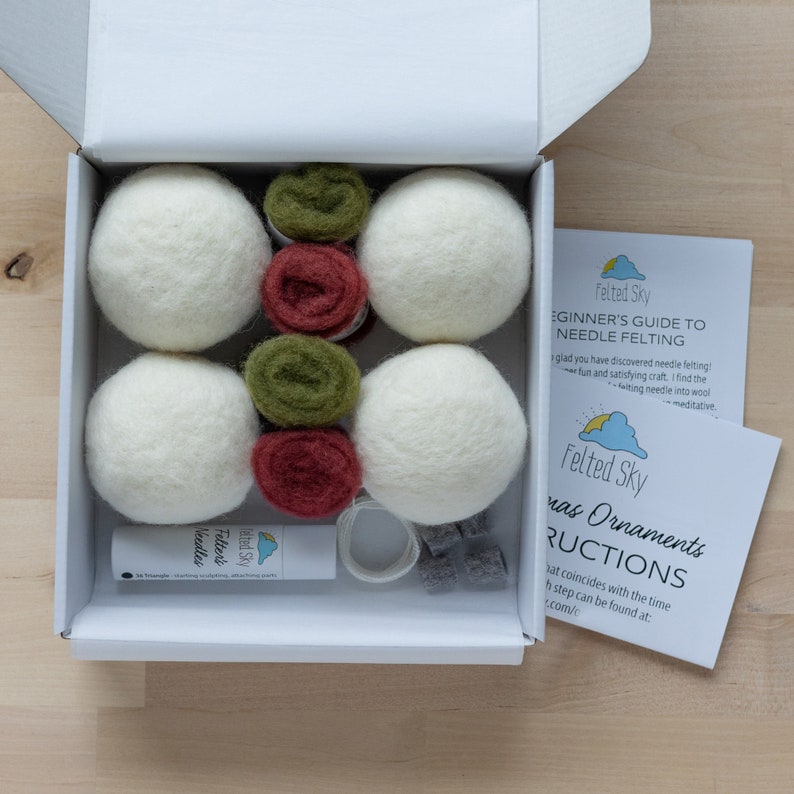 Op de afbeelding: Een witte doos met een wit deksel dat open is, waardoor een naaldviltset voor het maken van kerstversiering zichtbaar wordt. De set bevat witte wollen ballen, groene en rode viltwol, een naaldviltgereedschap en instructies. De doos staat op een houten oppervlak.