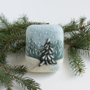 Op de afbeelding: Een blauwe en witte vilten ornament met een besneeuwde bos scène. Een enkele dennenboom staat op de voorgrond met een witte besneeuwde basis. De achtergrond is een blauwe lucht met witte sneeuwvlokken.