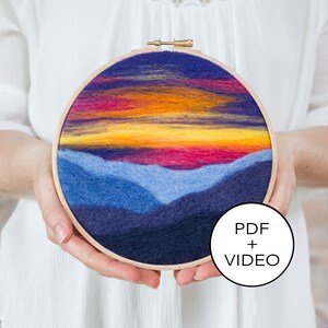 Op de afbeelding: Een naaldgevoelde zonsonderganglandschap met blauwe bergen en een gele, oranje en roze lucht in een houten borduurring. PDF- en video-instructies zijn inbegrepen.