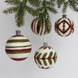 Op de afbeelding: Drie handgemaakte kerstballen van vilt met verschillende patronen. De ballen zijn wit met rode en groene accenten. De ballen hangen aan een dennentak.