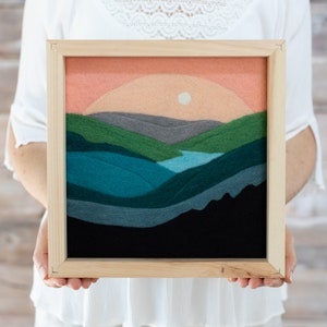 May include: A framed felt wall art piece depicting a landscape with a sunset over a lake and mountains. The mountains are layered in shades of green, blue, and gray, with a small lake in the center. The sunset is a soft orange and pink, with a white sun.