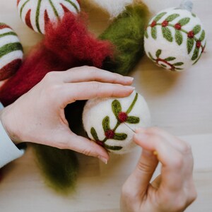 Op de afbeelding: Een persoon maakt een kerstbal van vilt. De bal is wit met groene bladeren en rode bessen. De persoon gebruikt een naald om de bladeren en bessen aan de bal te bevestigen.