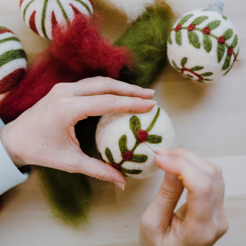Op de afbeelding: Een persoon maakt een kerstbal van vilt. De bal is wit met groene bladeren en rode bessen. De persoon gebruikt een naald om de bladeren en bessen aan de bal te bevestigen.
