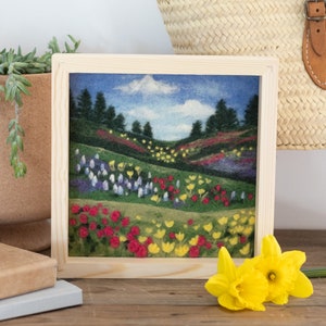 May include: A framed needle felted artwork depicting a colorful landscape with rolling hills, green grass, and a variety of flowers in shades of red, yellow, blue, and purple. The artwork is set against a blue sky with a single white cloud.