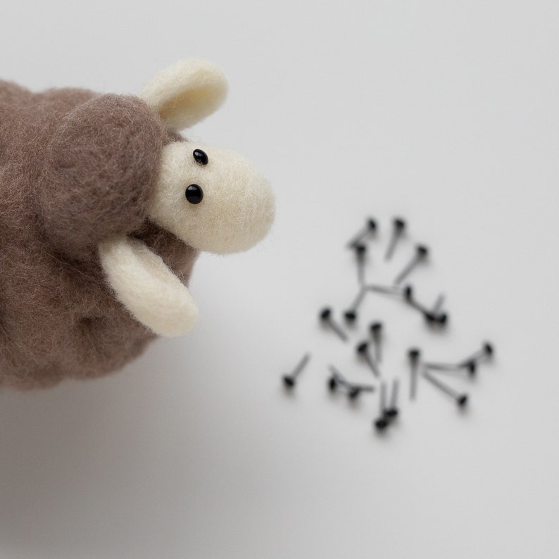 May include: A brown and white felt sheep toy with black eyes looking at a pile of black safety pins on a white surface.